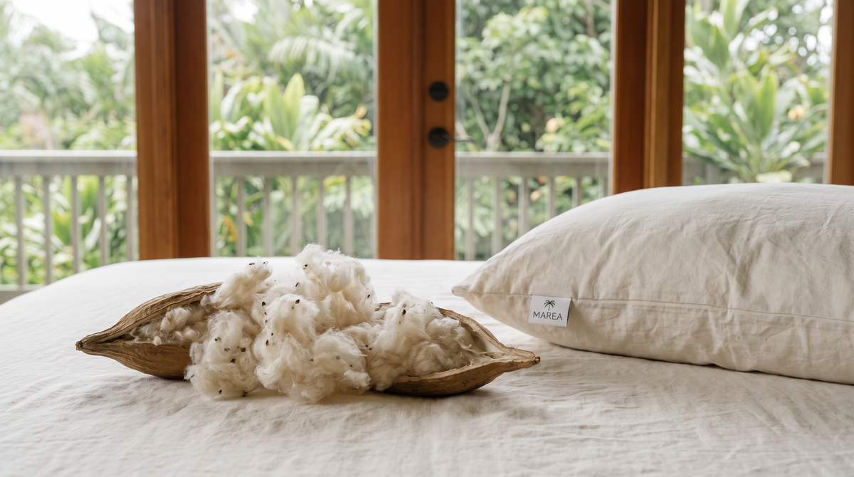 A split kapok seed pod spilling ecru fiber onto linen bedding beside a MAREA kapok pillow