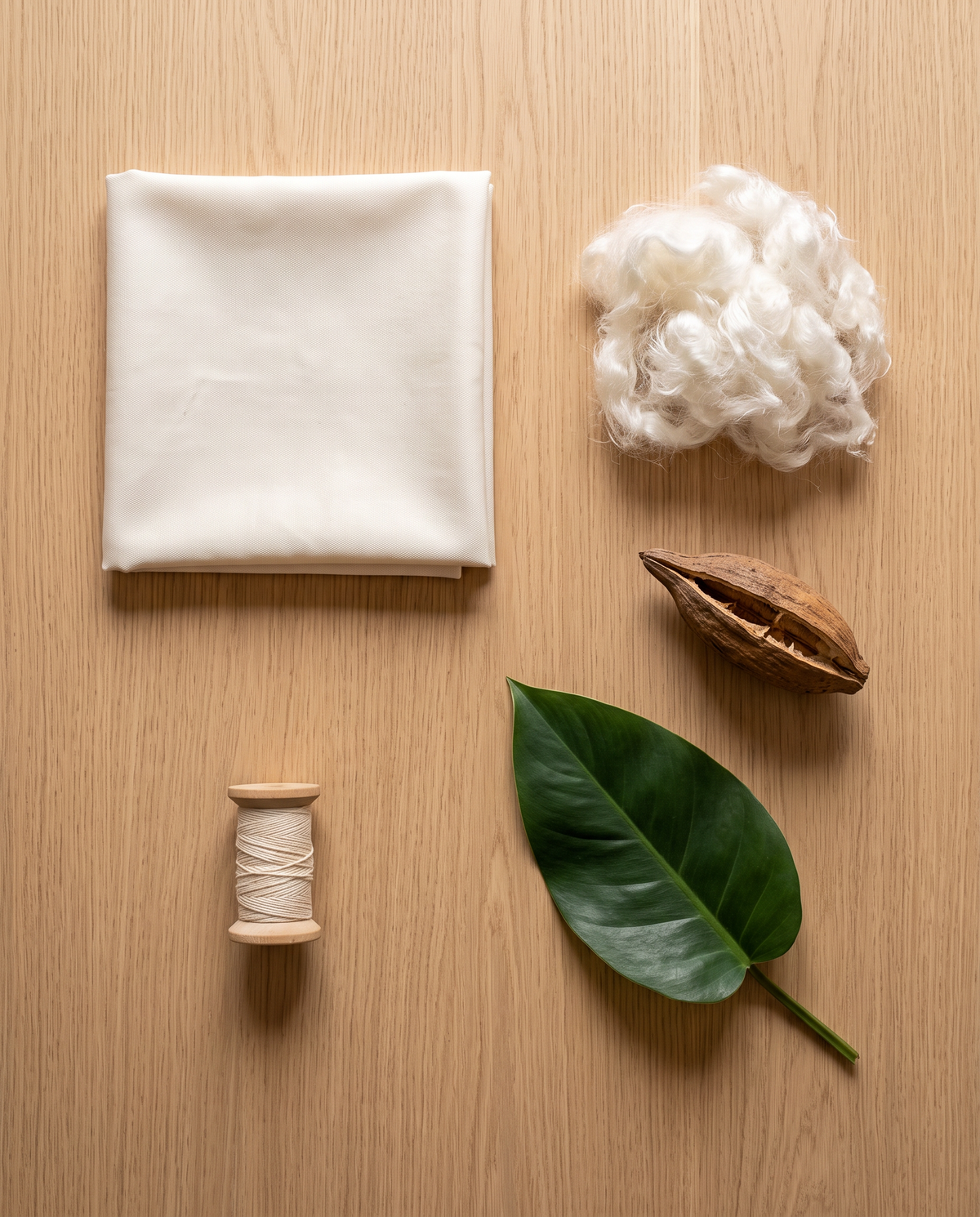 Flat lay of organic cotton sateen, kapok fiber, a kapok seed pod, cotton thread and a leaf on oak